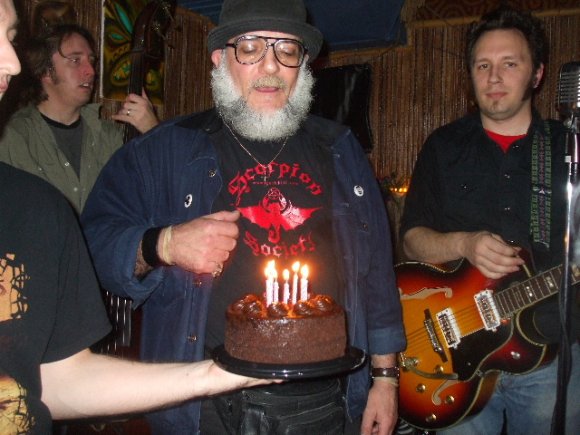 FRANKIE the KID holds the cake as Mr. WOOD prepares to blow out the candles as CHET & JORGE BEEFALO look on.